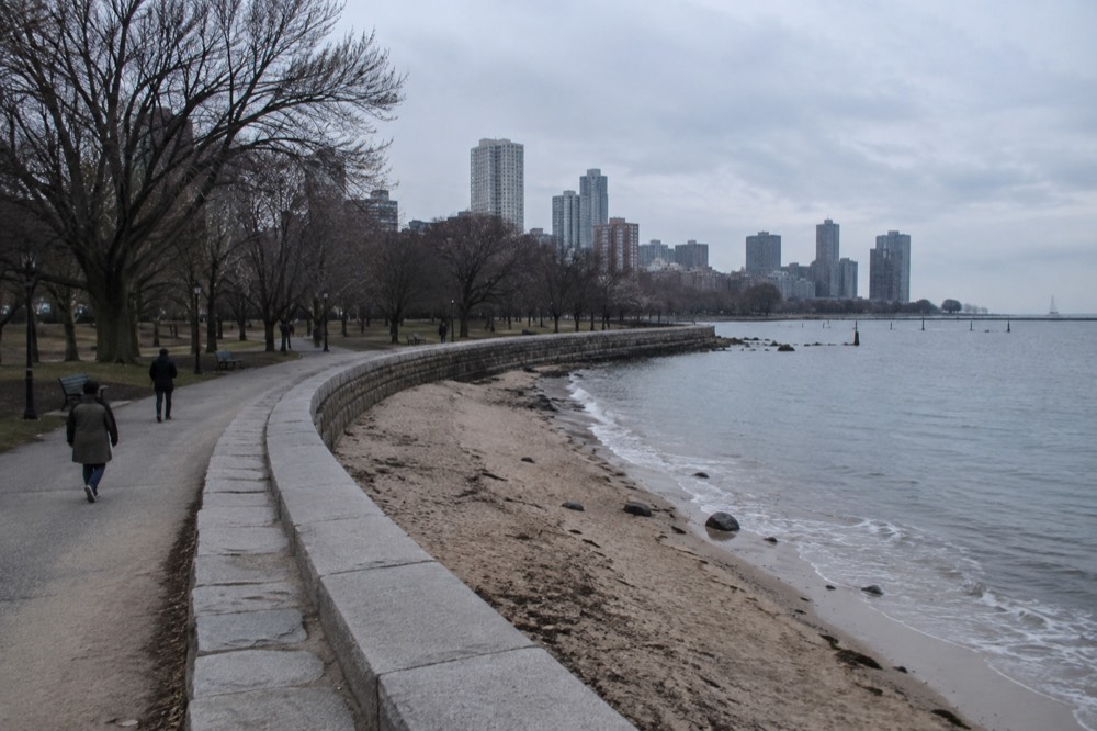 A sandy beach along Boston Harbor with a curved seawall and city skyline in the distance