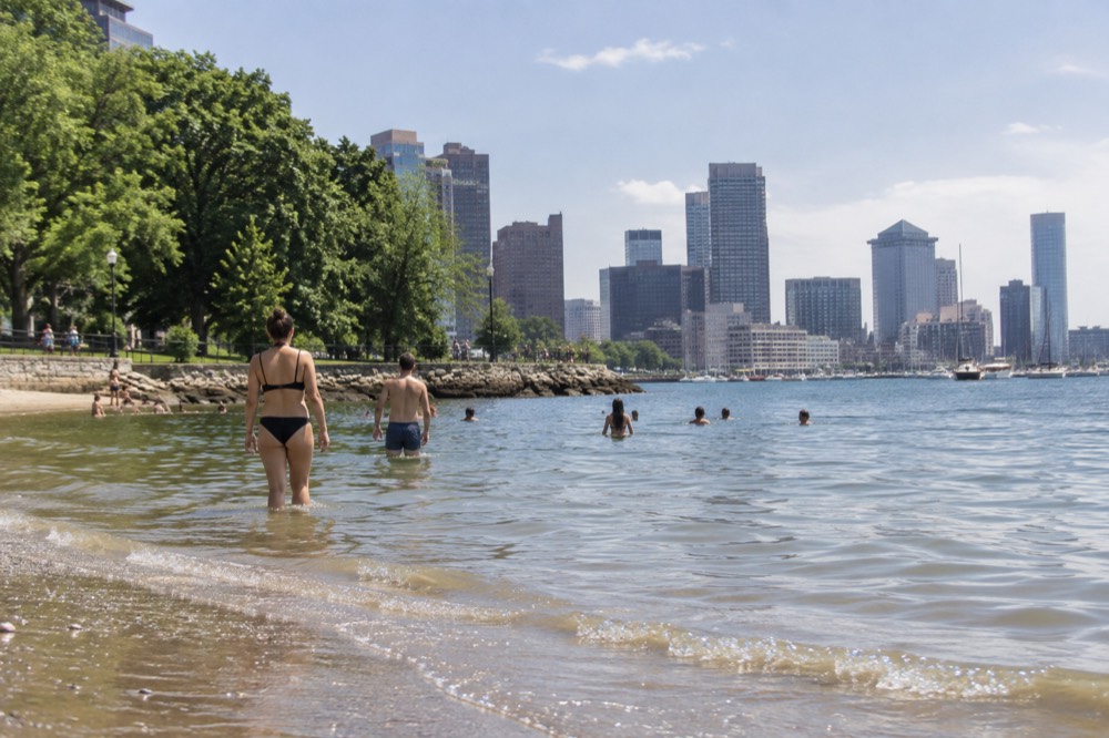 People wading into calm harbor water at a small city beach on a sunny summer afternoon