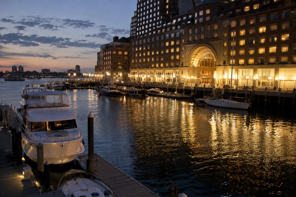An evening view of a lit hotel facade across a harbor wharf with moored boats