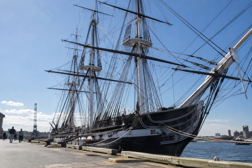 The USS Constitution moored in the Charlestown Navy Yard with masts rigged against a clear sky
