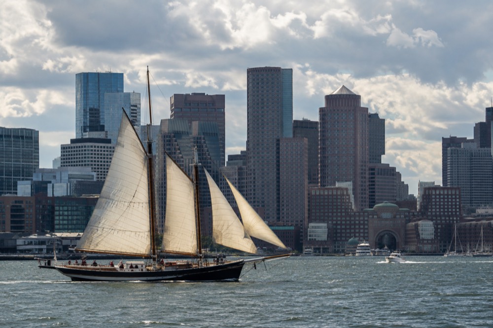 A tall ship with sails up crossing Boston Harbor with the downtown skyline behind it