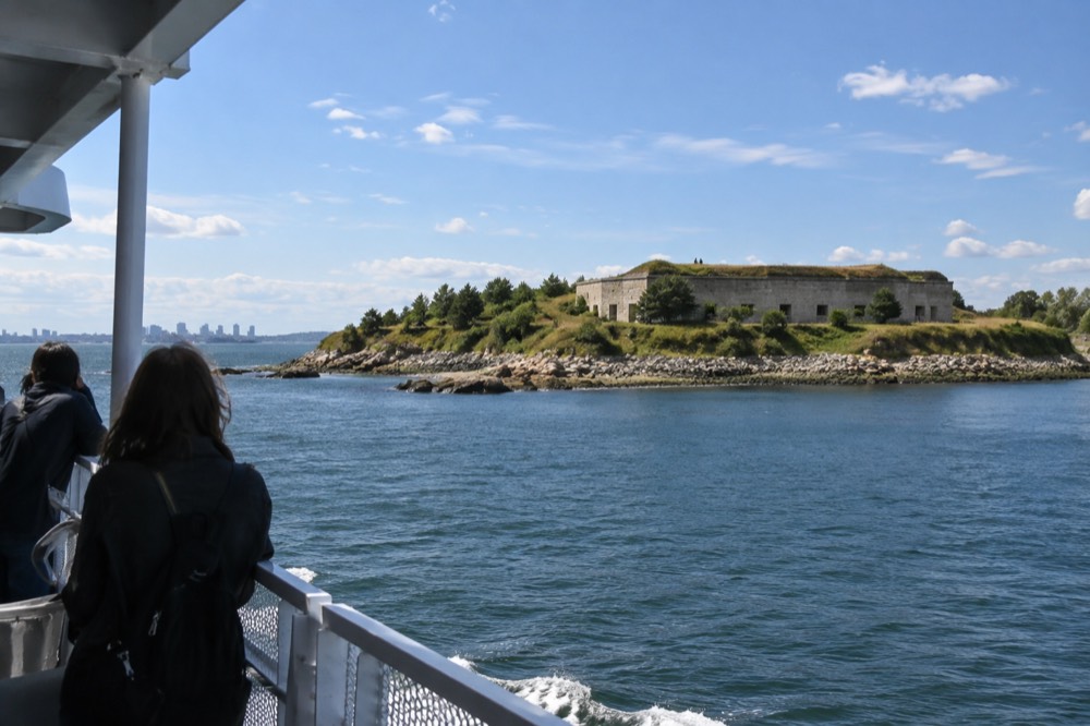 A ferry approaching a green harbor island with a granite fort under a bright sky