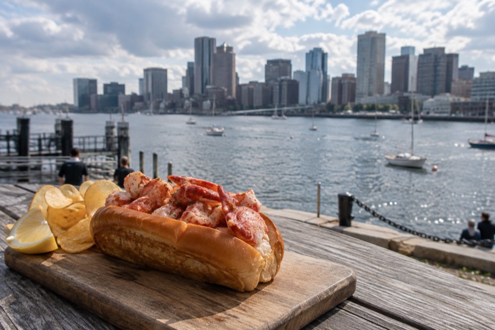 A lobster roll in a buttered split-top bun on a wooden board beside a harbor view