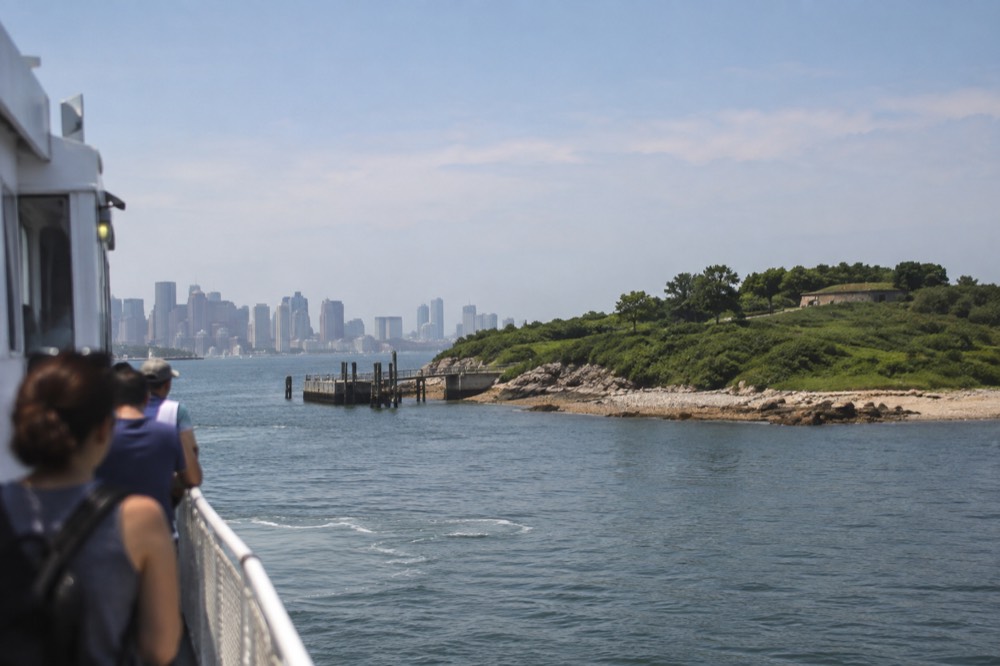 A ferry approaching a green harbor island with a downtown Boston skyline in the distance