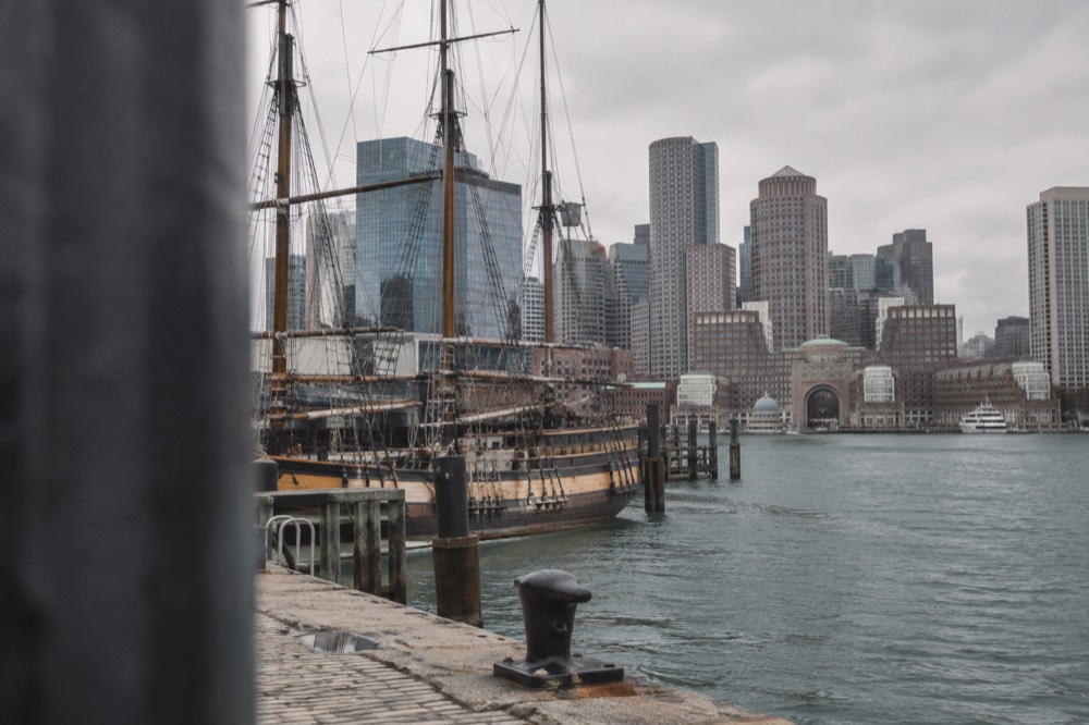 A wooden tall ship moored in Boston Harbor with downtown buildings behind it