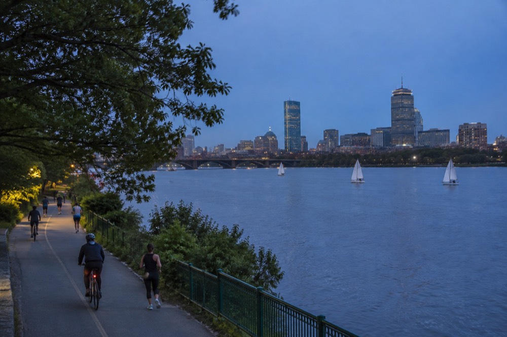 Runners and cyclists on the Charles River Esplanade path with sailboats on the river