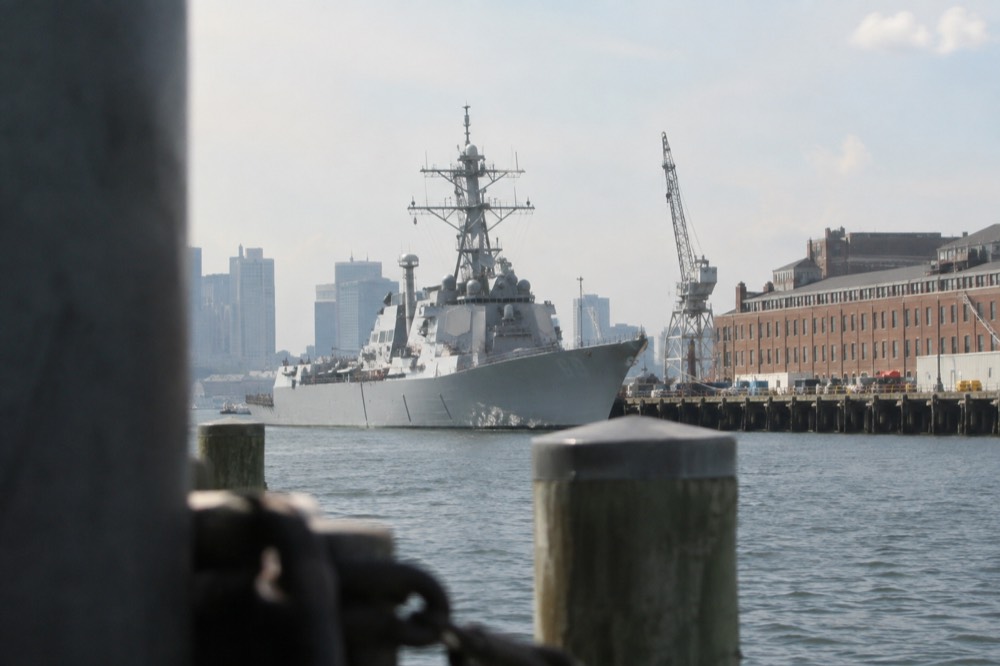 Gray warship moored at a pier with historic brick shipyard buildings along the waterfront