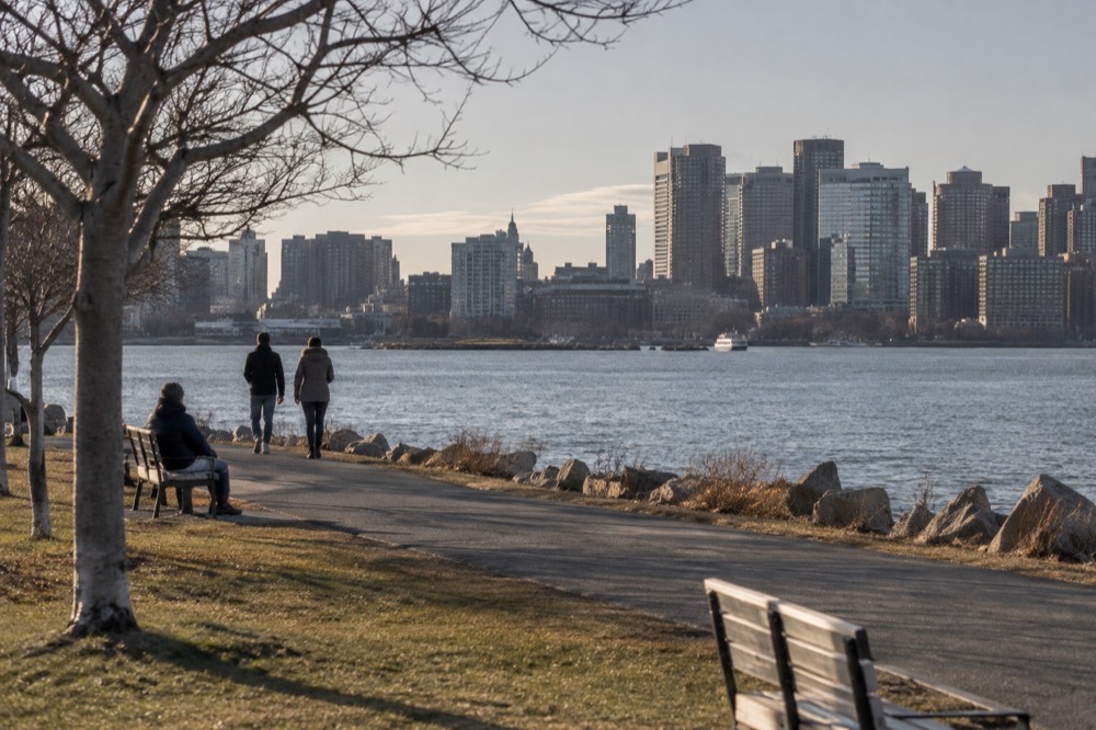A harbor shoreline path with grass, benches and a view across the water to the city skyline