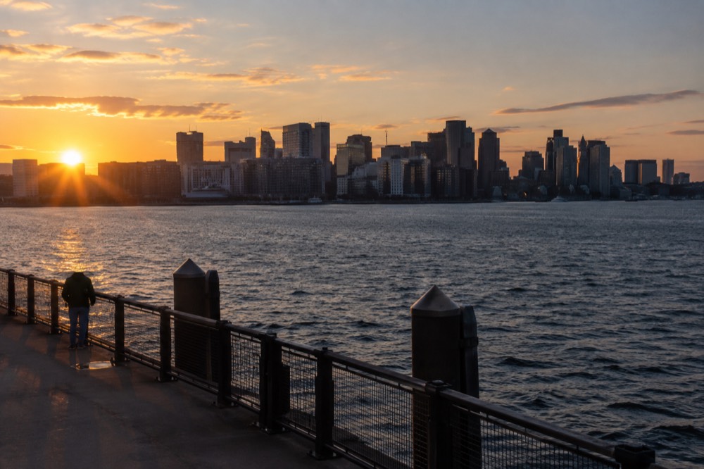 A city skyline across dark water at sunset with a pier railing in the foreground