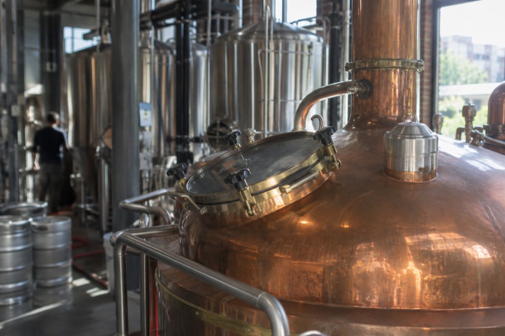 A copper brewing kettle inside a brewery with stainless tanks behind it