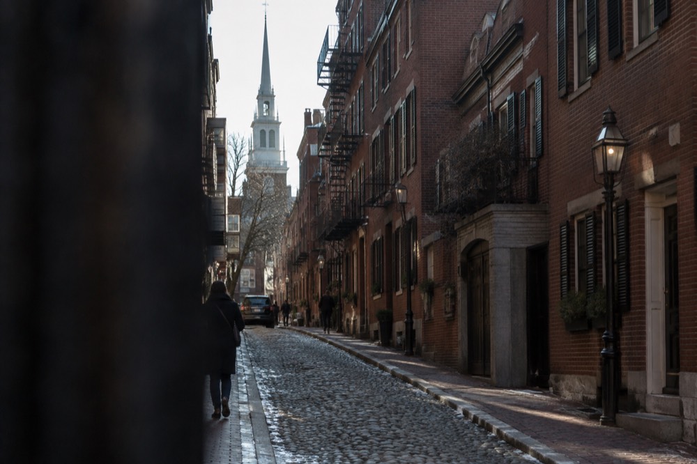 Narrow cobblestone street in the North End with brick row houses and an old church spire