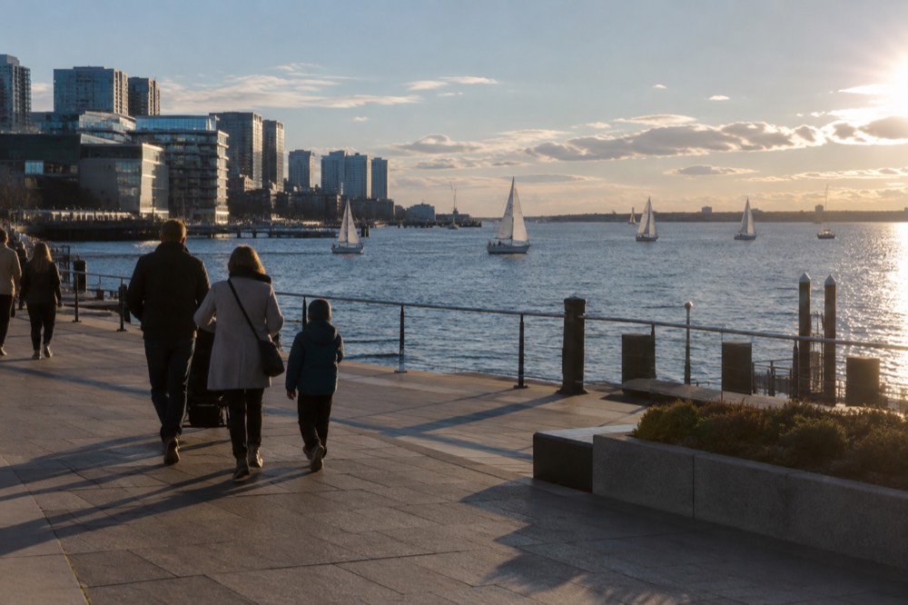 A family walking along a wide waterfront promenade with sailboats on Boston Harbor behind them