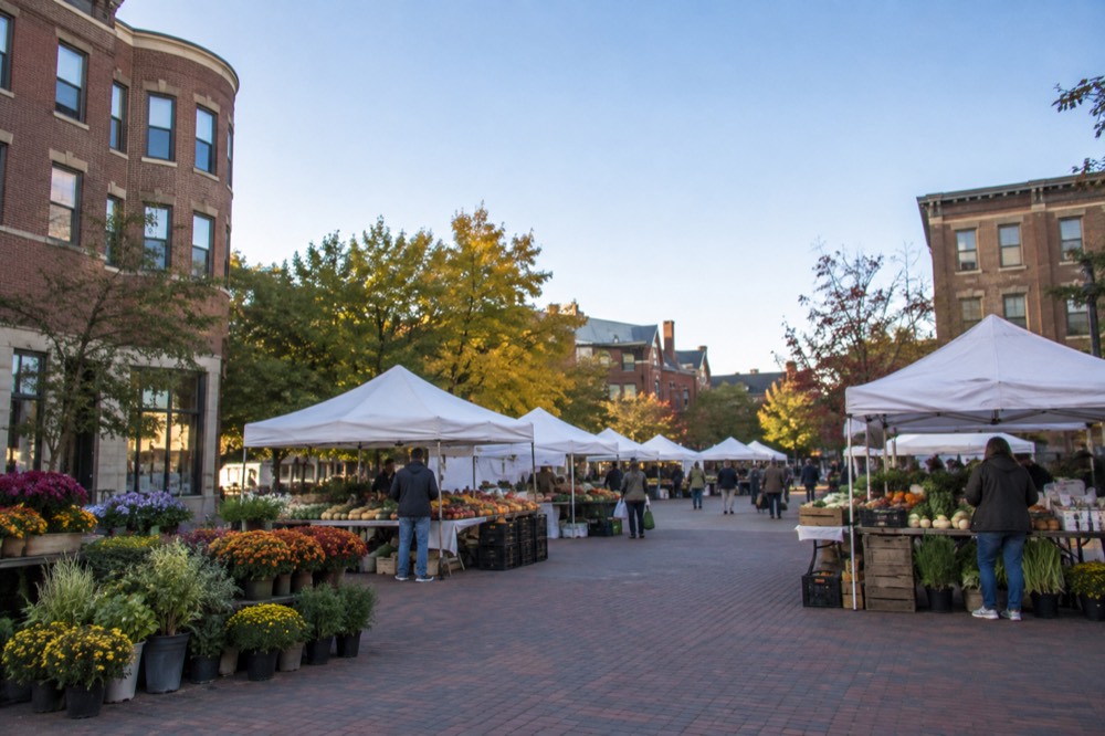 A neighborhood square with a farmers market, white tents, and brick buildings on a sunny morning
