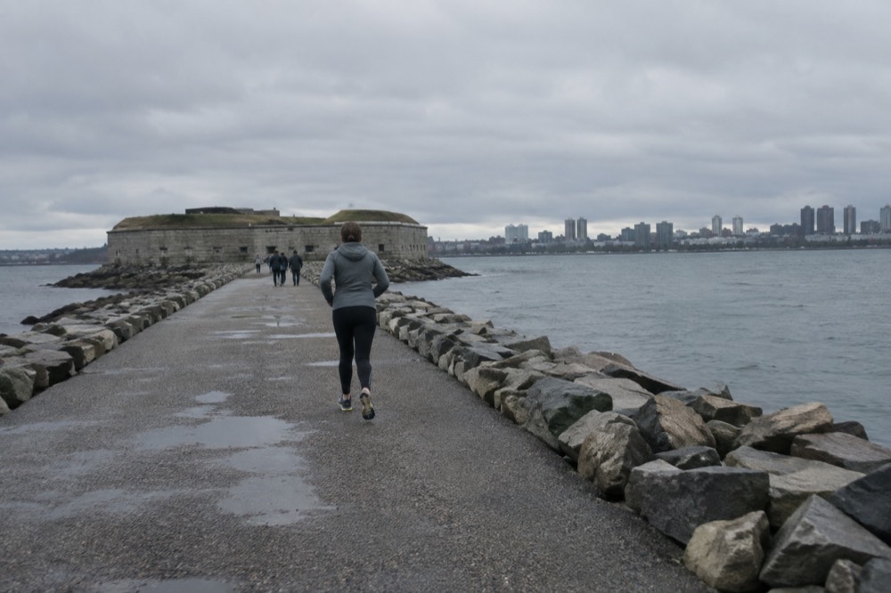 A stone fortification on a small island connected to a causeway, with a harbor and city skyline in the distance