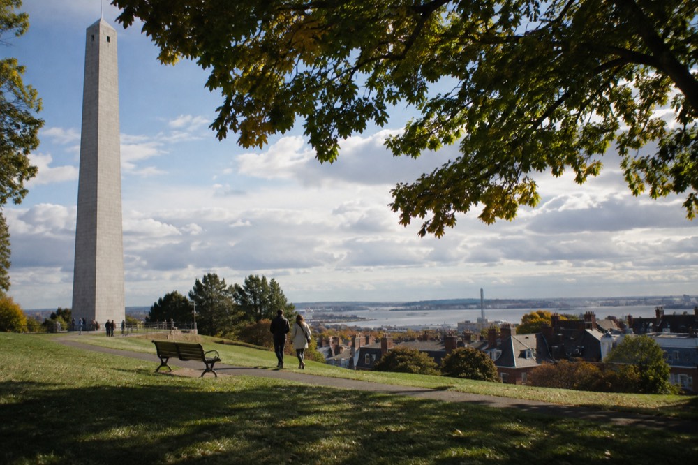 A grassy hilltop park with a white stone monument tower and views over rooftops toward a harbor