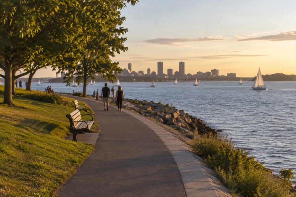 A curving beach promenade with a wide walkway, grass, and calm water with sailboats offshore