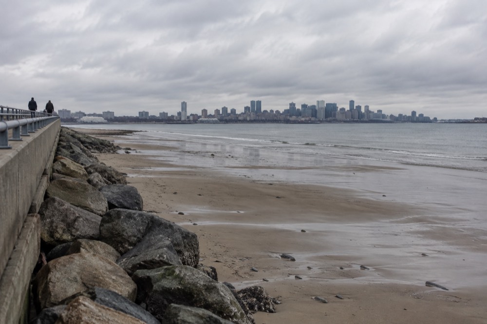 A sandy beach at low tide with exposed wet sand, a long walkway, and a city skyline across the water