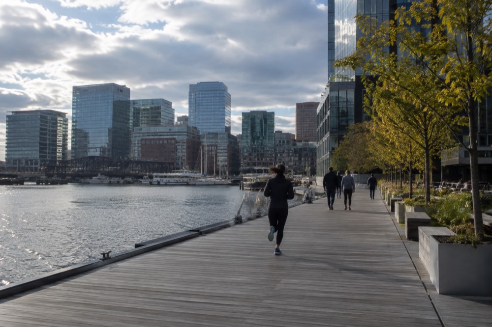 Modern glass buildings along a waterfront boardwalk with small trees and people walking
