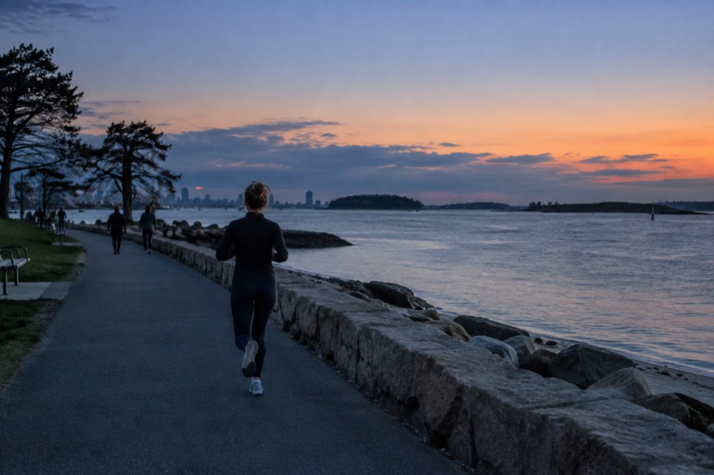 A harbor shoreline at sunset with calm water, distant islands, and a low stone seawall