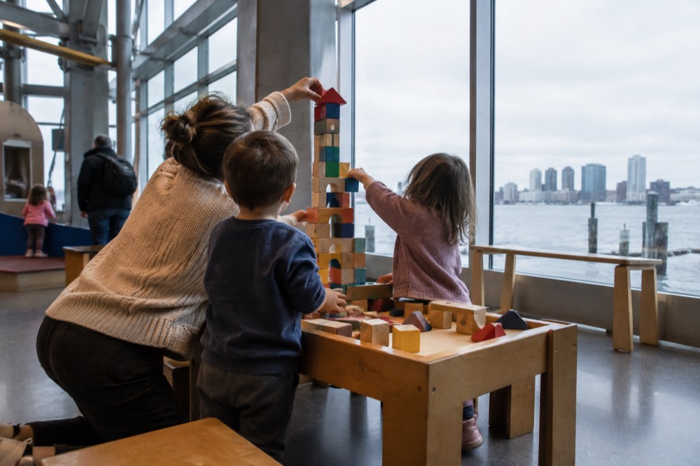 A parent and two children building a tall block tower together at a low museum table