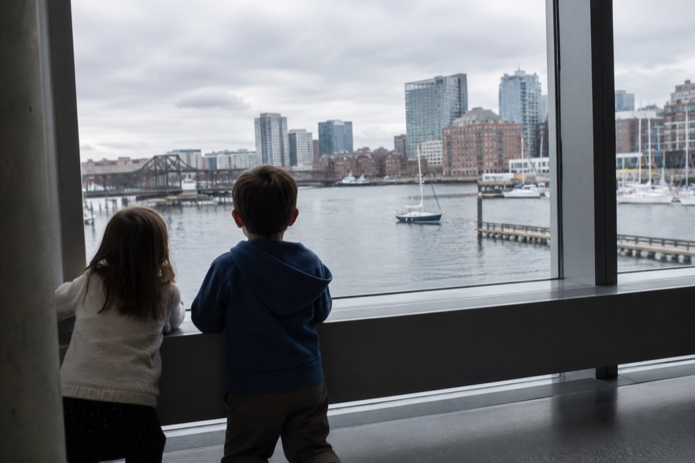 Two children looking out a large museum window toward boats on a city waterway