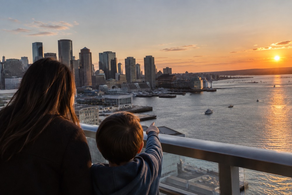 A city skyline seen at sunset from a high observation deck with a child pointing toward the harbor