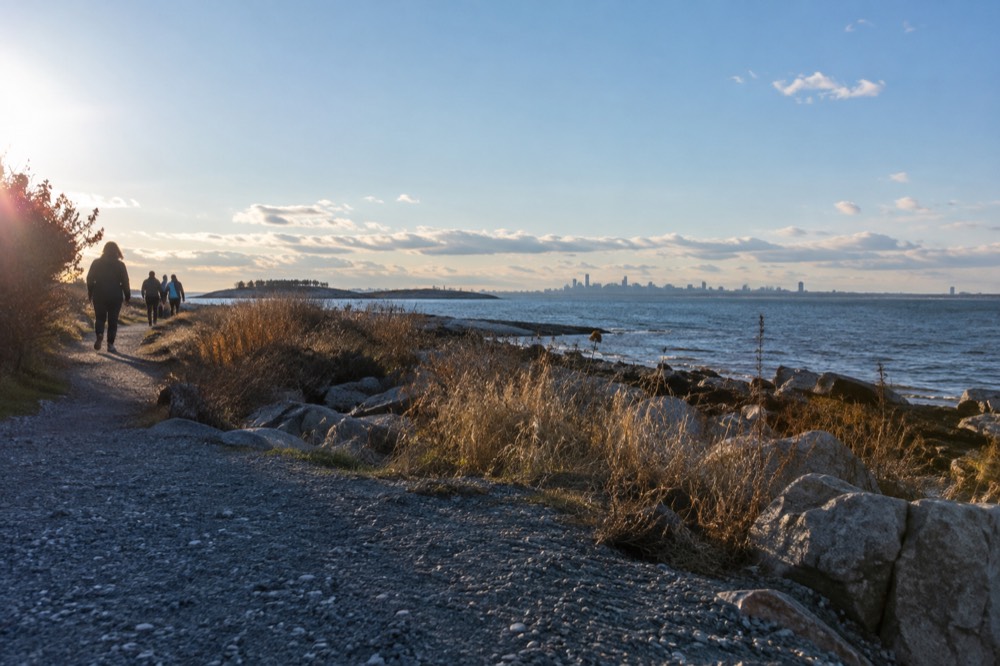 Visitors walking a gravel trail toward a harbor island shoreline under a blue sky