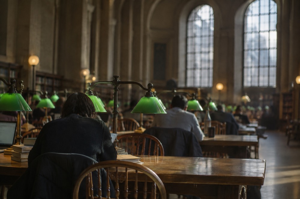 Grand reading room with tall arched windows and rows of green reading lamps over long wooden tables