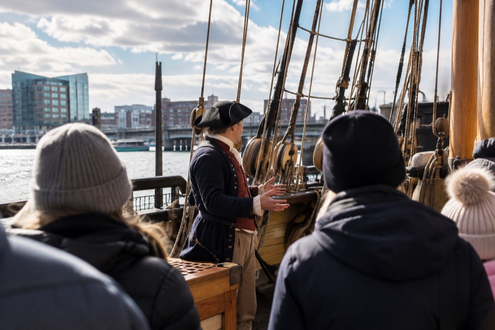 Costumed interpreter in colonial dress speaking to visitors on a wooden ship deck