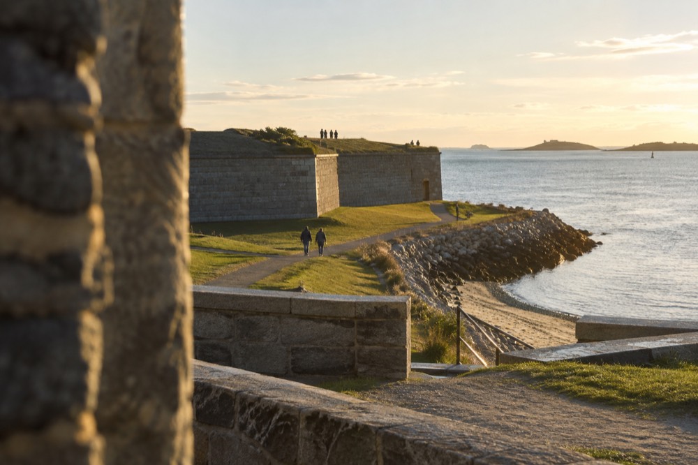 A granite fort at golden hour with calm harbor water and distant islands on the horizon