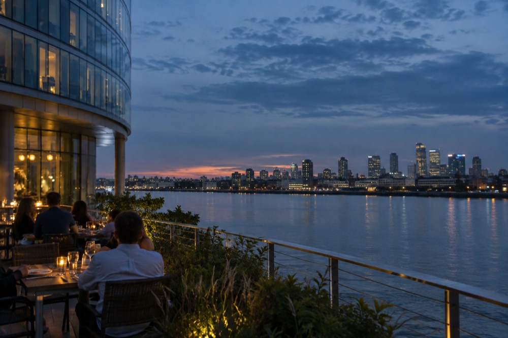 Diners at an outdoor waterfront table with the Boston skyline across the river