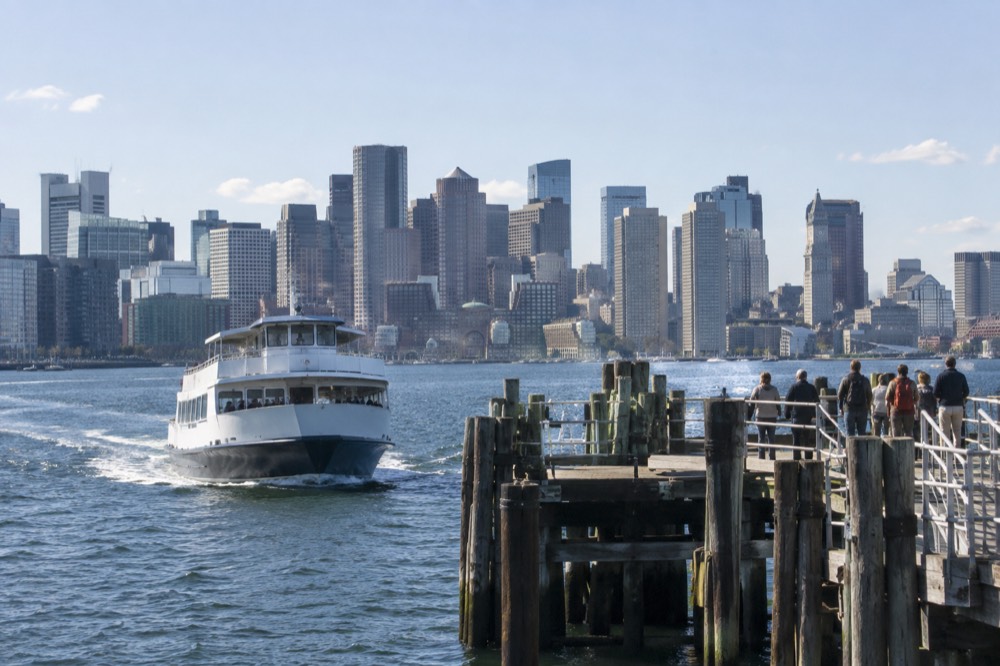 A harbor ferry approaching a wooden island pier with the Boston skyline across the water
