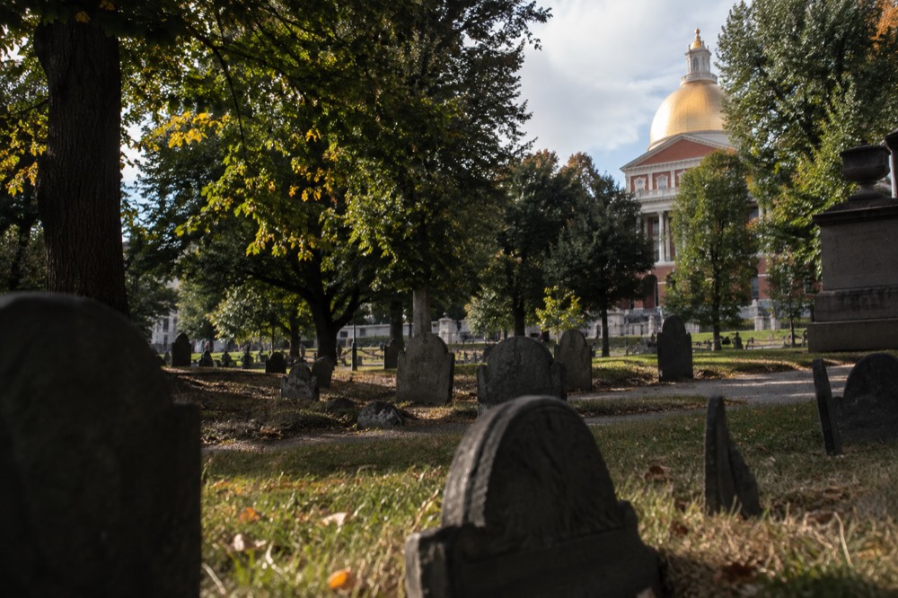 The golden dome of the Massachusetts State House visible above trees from a Boston cemetery