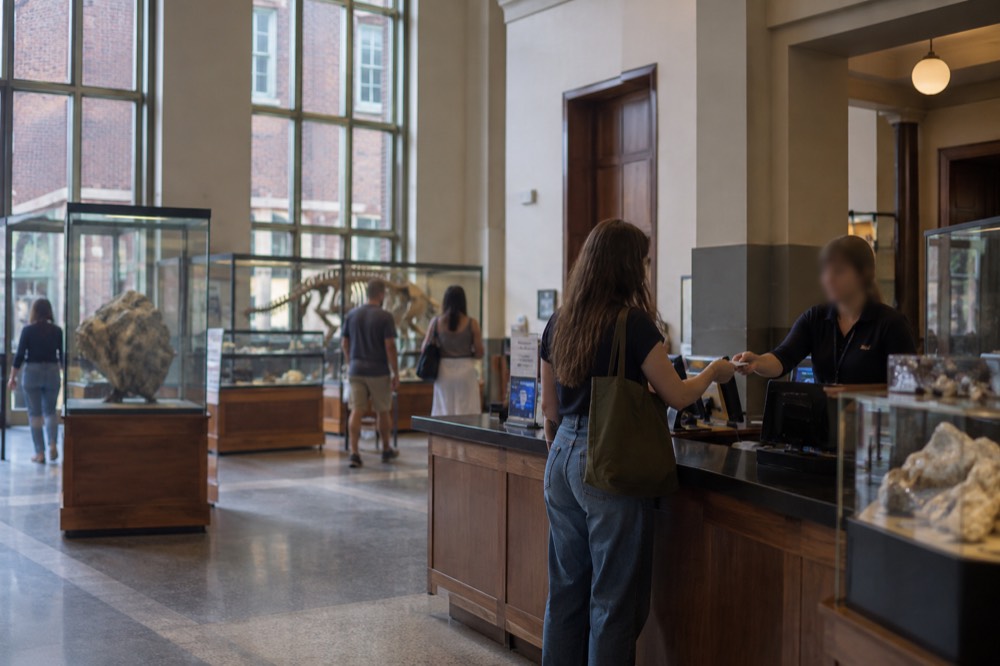A museum ticket counter with a visitor paying while staff hand over printed admission tickets