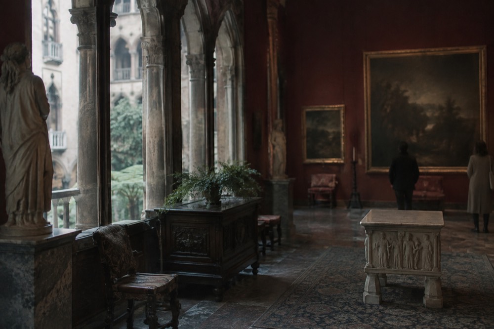 A gallery interior with tall windows, stone columns and paintings on dark red walls