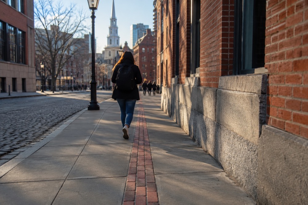 A red brick Freedom Trail line embedded in a Boston sidewalk leading toward historic buildings