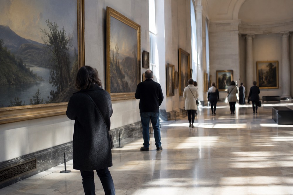 A museum gallery with visitors viewing large framed paintings in a bright hall