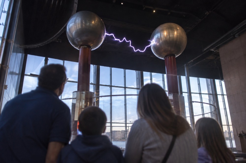 A family watching a large indoor electricity demonstration between two metal spheres