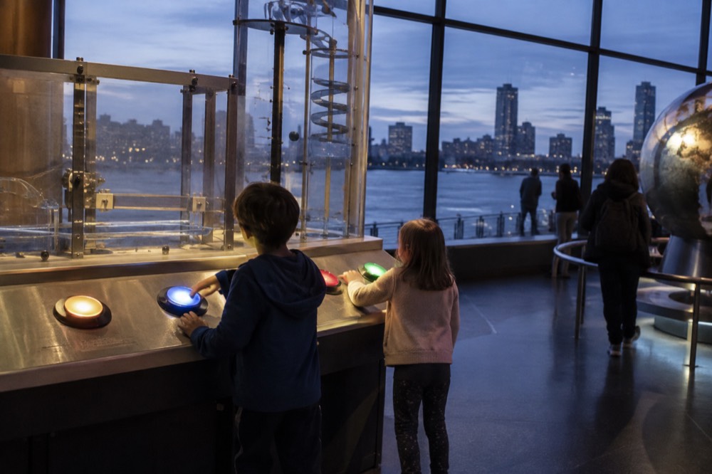Children pressing buttons at a hands-on science exhibit table inside a bright museum hall