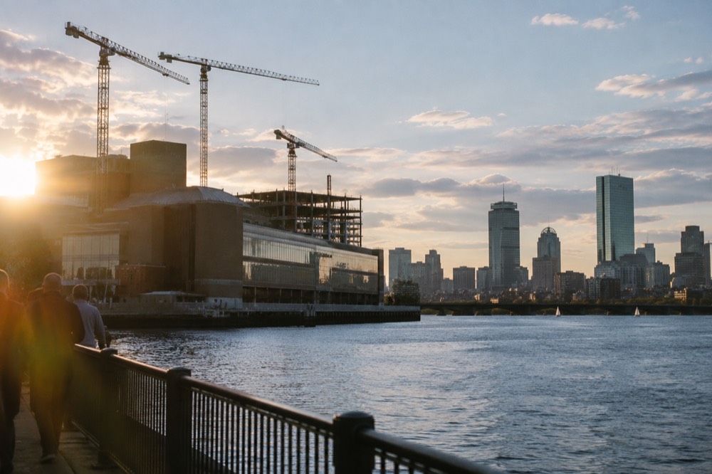 Construction cranes above a riverside museum building with the Boston skyline behind