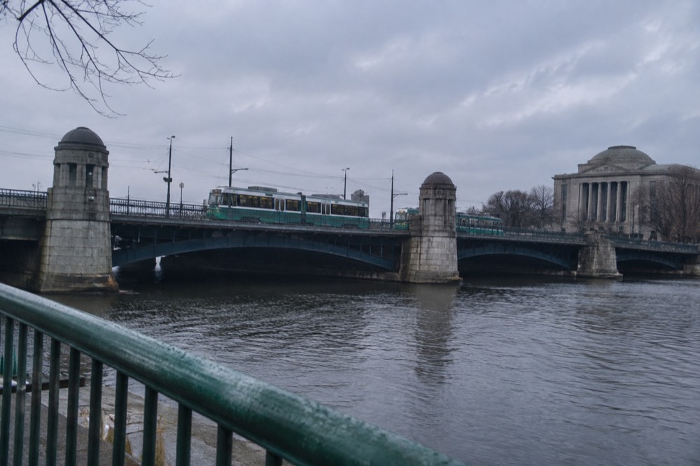 A Green Line trolley crossing an iron bridge over the Charles River near a museum building