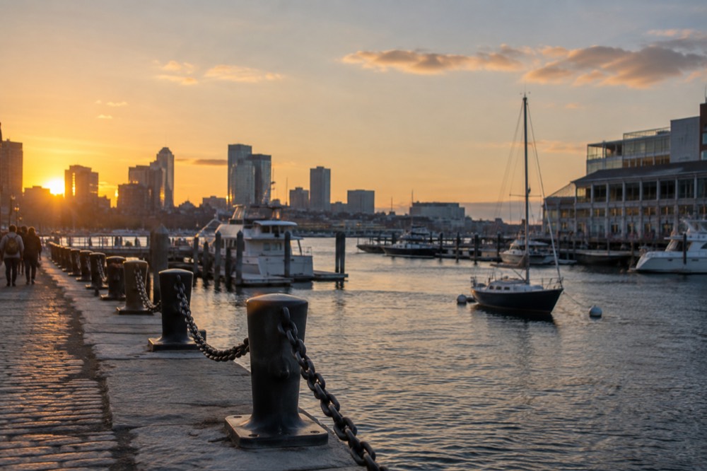 A harborfront walkway with moored boats, a wooden pier, and city buildings along the shoreline