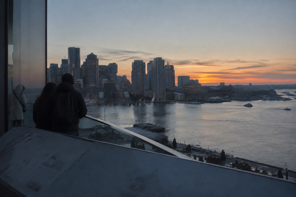 A city skyline seen from a high observation deck at dusk with harbor water in the distance