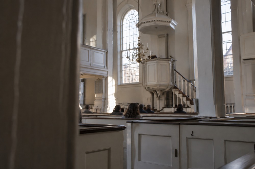 Interior of a colonial-era church with white box pews and a raised pulpit under a chandelier