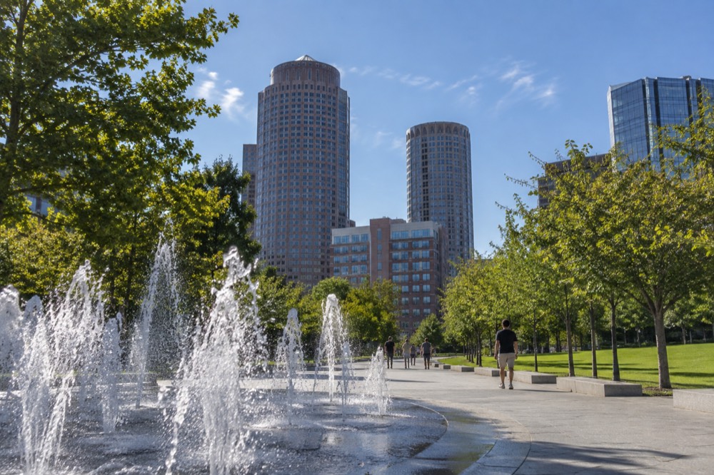 A tree-lined park path with fountains, lawn and city towers behind it on a sunny day
