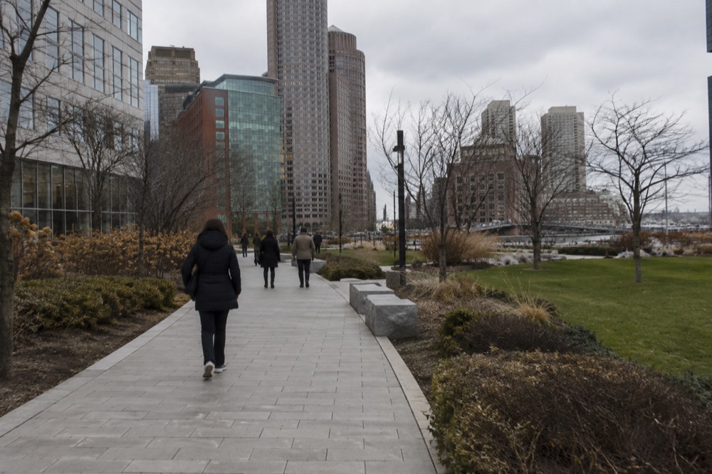 A green park corridor between office towers with people walking a wide stone path