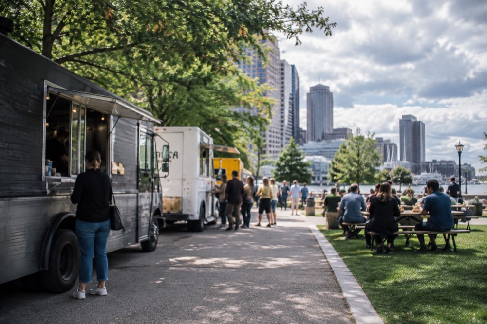 Food trucks parked along a park path with people eating at picnic tables