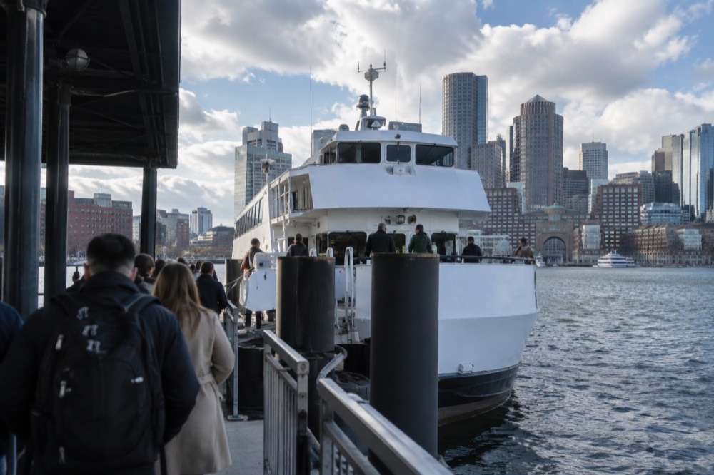 Passengers boarding a white ferry at a downtown Boston pier with a skyline behind it