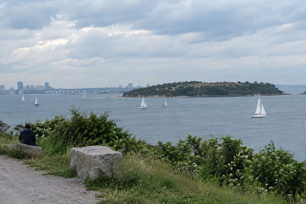 A wide harbor view with sailboats and a green island on the horizon under scattered clouds