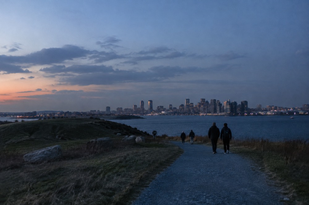 Visitors in jackets walking a gravel trail near a hilltop with the Boston skyline in the distance